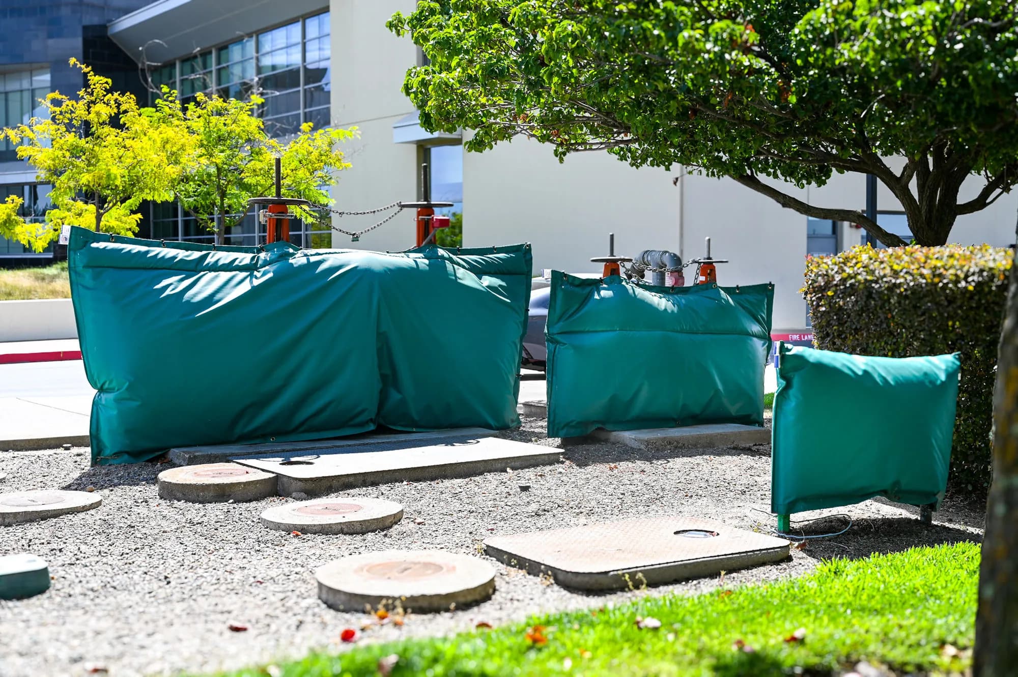 Three green insulated backflow covers in a gravel bed outside an office building — a large double-check detector assembly, a mid-size assembly, and a small companion cover.