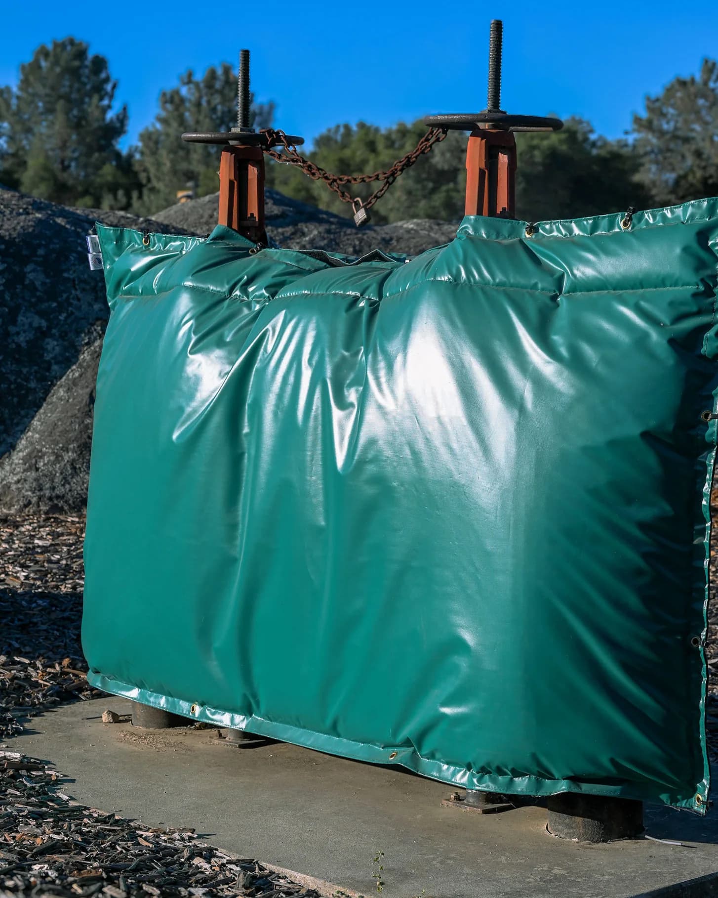 Green insulated backflow cover installed against a granite outcrop under a clear blue sky, with a padlocked chain and orange valve stems rising above the cover.