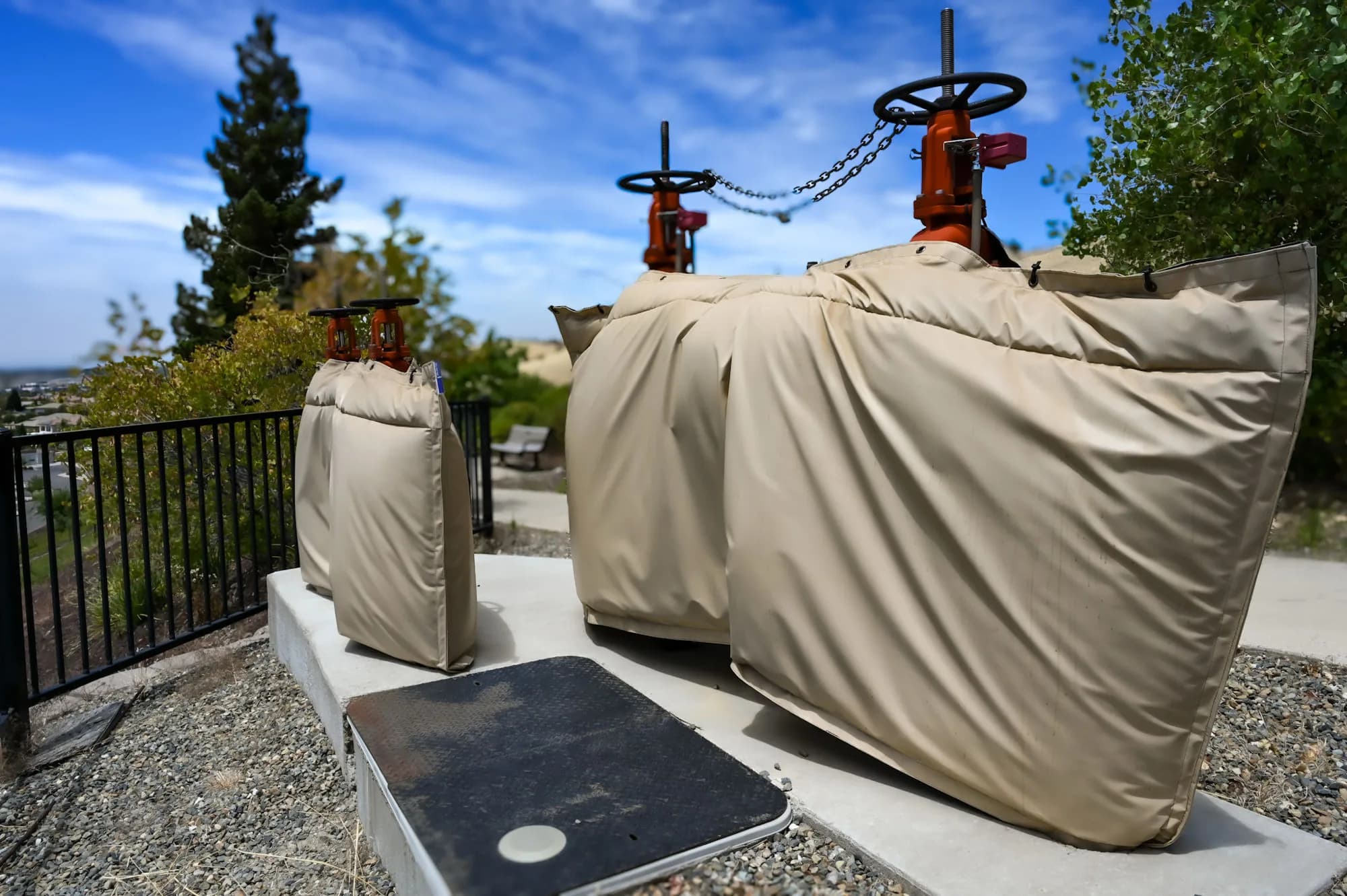 Two tan insulated backflow covers on a concrete pad above a hillside neighborhood — a full-size assembly cover beside a smaller companion cover, with the red gate valves left exposed for access.