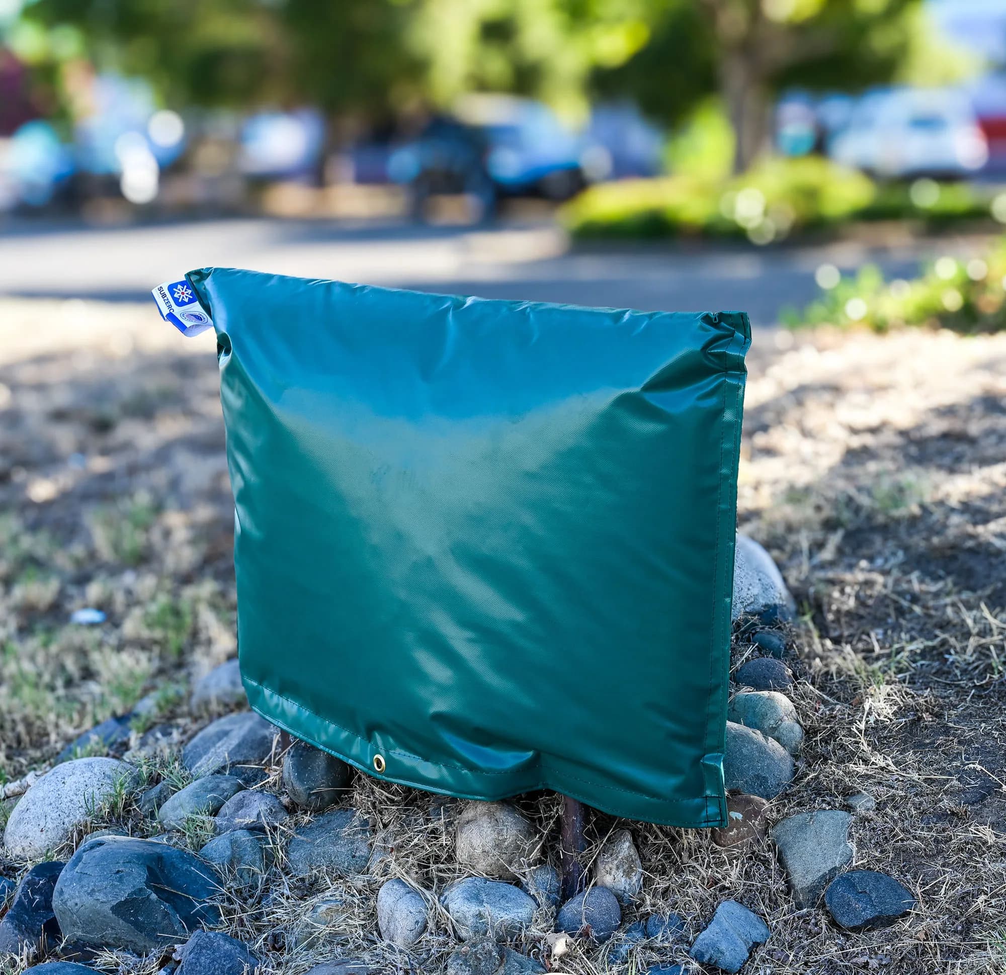 Small green insulated backflow cover seated on river-rock landscaping beside a road, carrying the Sub Zero Covers product tag.